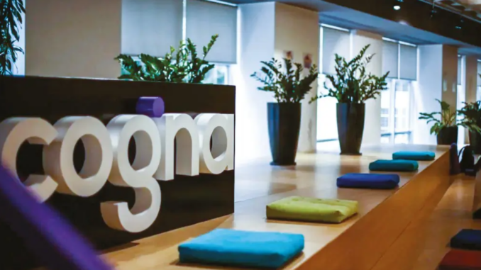 Logotipo da Cogna em área interna corporativa com plantas e ambiente moderno ao fundo.