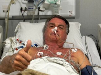 Jair Bolsonaro deitado em cama hospitalar com eletrodos e suporte médico, levantando o polegar em sinal de positivo