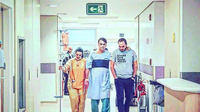 Jair Bolsonaro caminhando em corredor de hospital acompanhado por equipe de apoio, com acesso venoso e suporte médico ao lado.