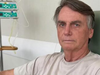 Jair Bolsonaro sentado em ambiente hospitalar, com acesso intravenoso ao fundo, durante tratamento médico.