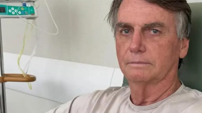 Jair Bolsonaro sentado em ambiente hospitalar, com acesso intravenoso ao fundo, durante tratamento médico.