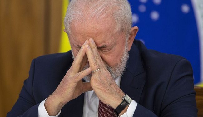 Presidente Luiz Inácio Lula da Silva com mãos unidas no rosto em expressão séria durante cerimônia oficial.