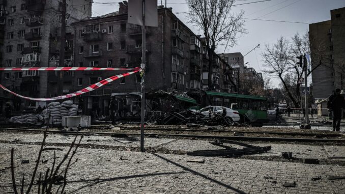 Rua destruída após explosão em área urbana, com prédio danificado, escombros, faixas de isolamento e veículos atingidos.