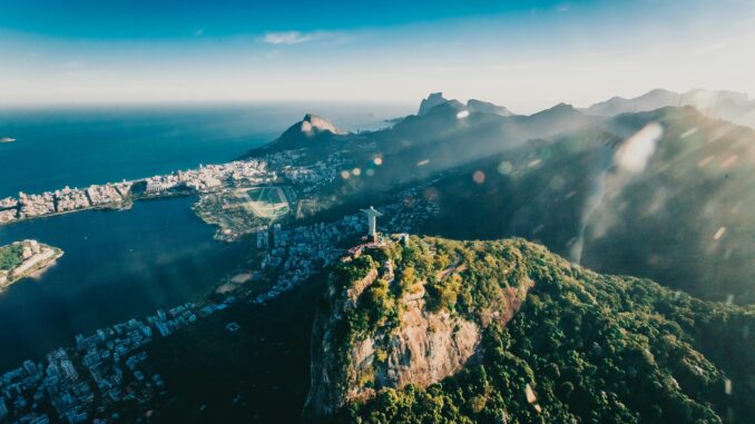 Vista aérea do Cristo Redentor no Rio de Janeiro, com montanhas, mar e cidade ao fundo em dia ensolarado.