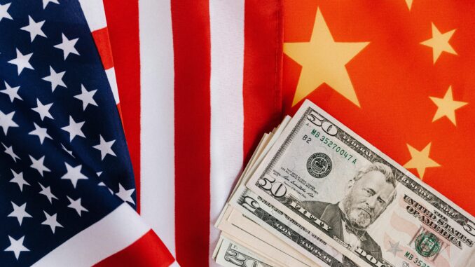 Bandeiras dos Estados Unidos e da China lado a lado com cédulas de dólares à frente, representando tensões econômicas e comerciais.