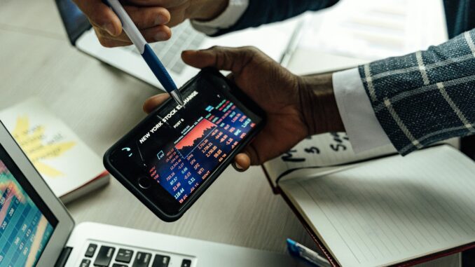 Investidor analisa dados da Bolsa de Nova York em smartphone, com gráficos de mercado e variações de preços em ambiente financeiro