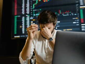 Homem sentado em frente ao notebook, segurando os óculos com expressão de estresse, com gráficos de mercado exibidos ao fundo