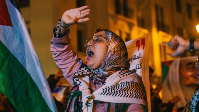 Mulher com hijab segurando bandeira e protestando em apoio à Palestina durante manifestação noturna.
