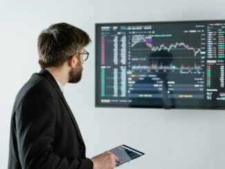 Homem analisando gráficos financeiros em painel digital enquanto segura tablet com dados de mercado